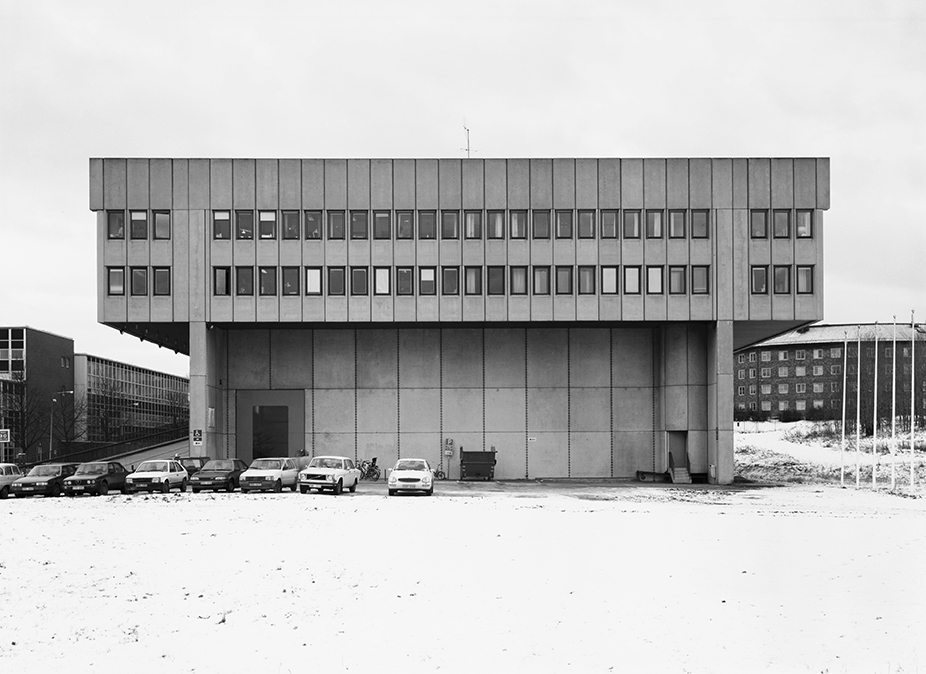 En rektangulär betongbyggnad med fönsterrader står på pelare ovanför snötäckt mark, med flera bilar parkerade framför.
