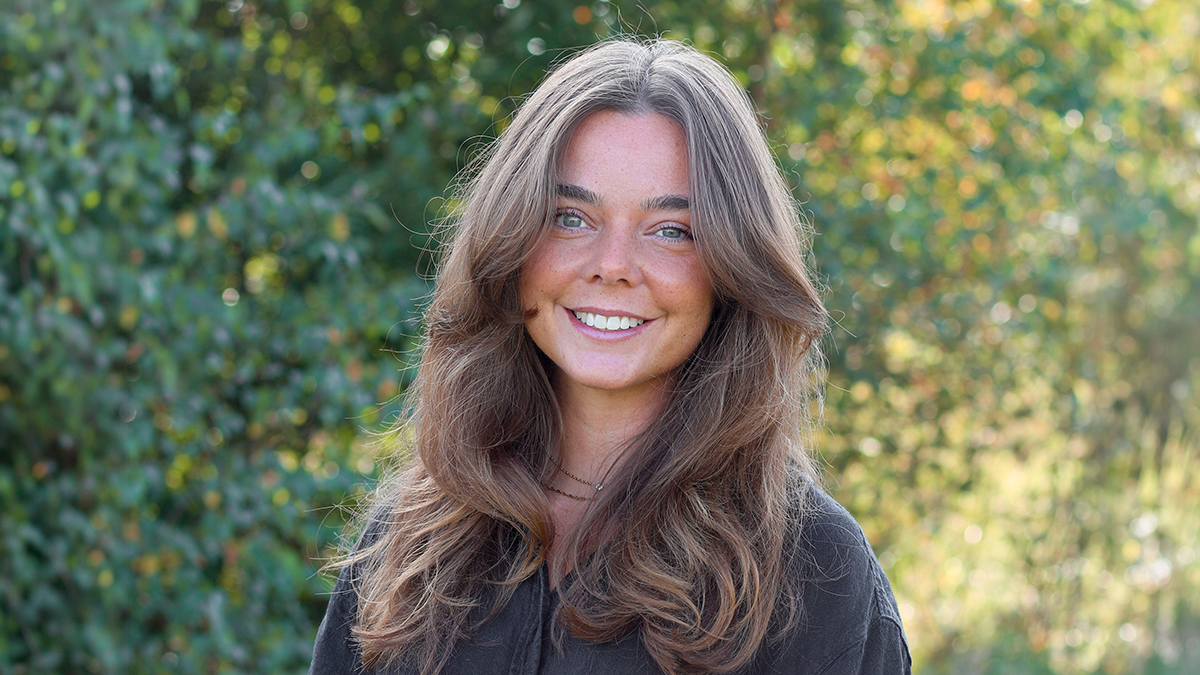 En kvinna med långt brunt hår och svart topp ler utomhus med växtlighet i bakgrunden.