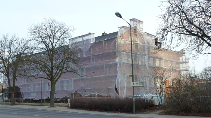 En tegelbyggnad täckt av byggnadsställningar och skyddsnät, med träd utan löv och en gatlampa i förgrunden.