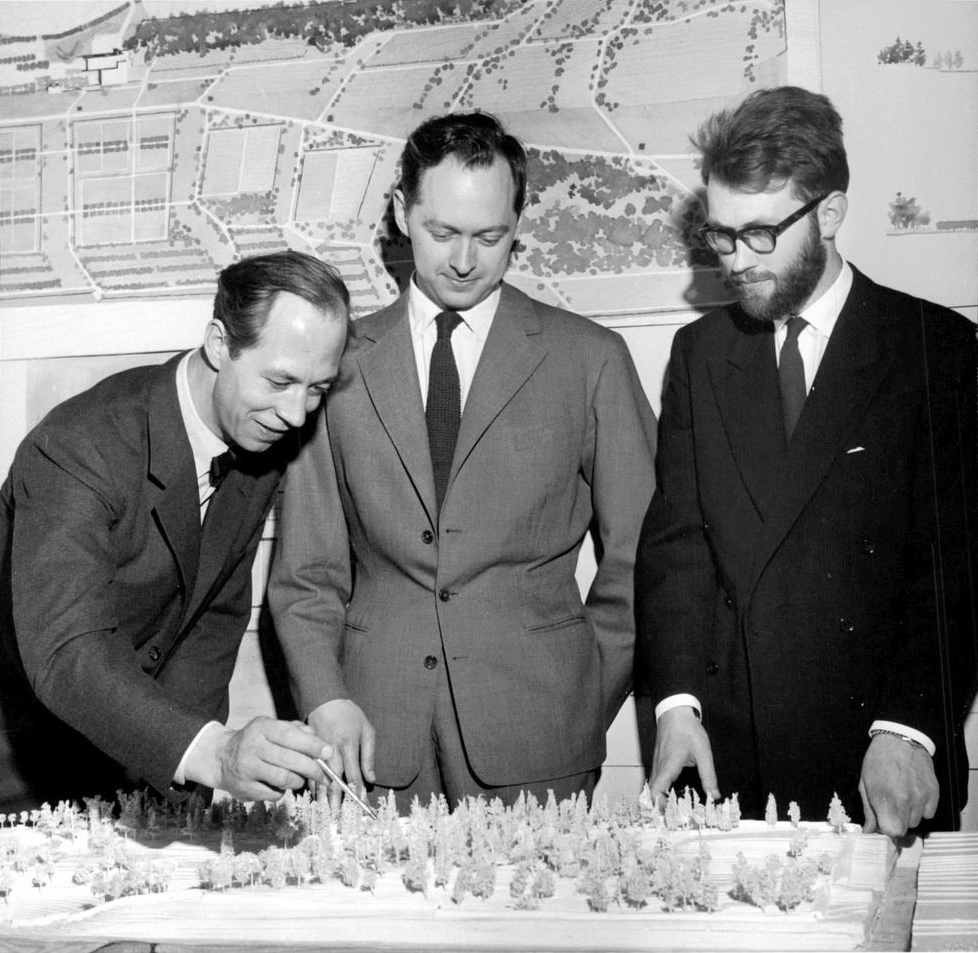 Tre män i kostymer tittar på och diskuterar en modell av ett landskapsprojekt, med en situationsplan på väggen bakom dem.