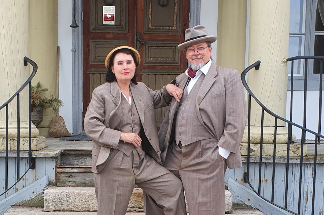 Två individer i vintagekläder poserar på trappor utanför en byggnad.