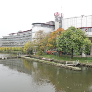 Modern byggnad med cylindriska och rektangulära sektioner vid en flod med en gångbro och höstiga träd.