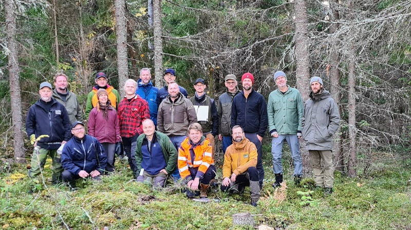 Grupp människor poserar i en skog, några stående och några på knä, iklädda utomhuskläder.