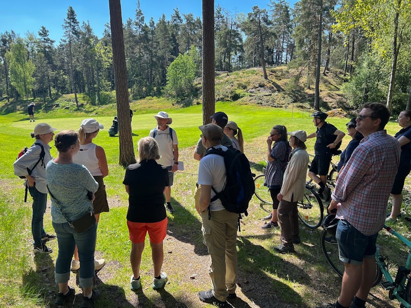 En grupp människor står i ett skogsområde och lyssnar på Micke Madenteg som berättar om Björkhagens golfklubb.