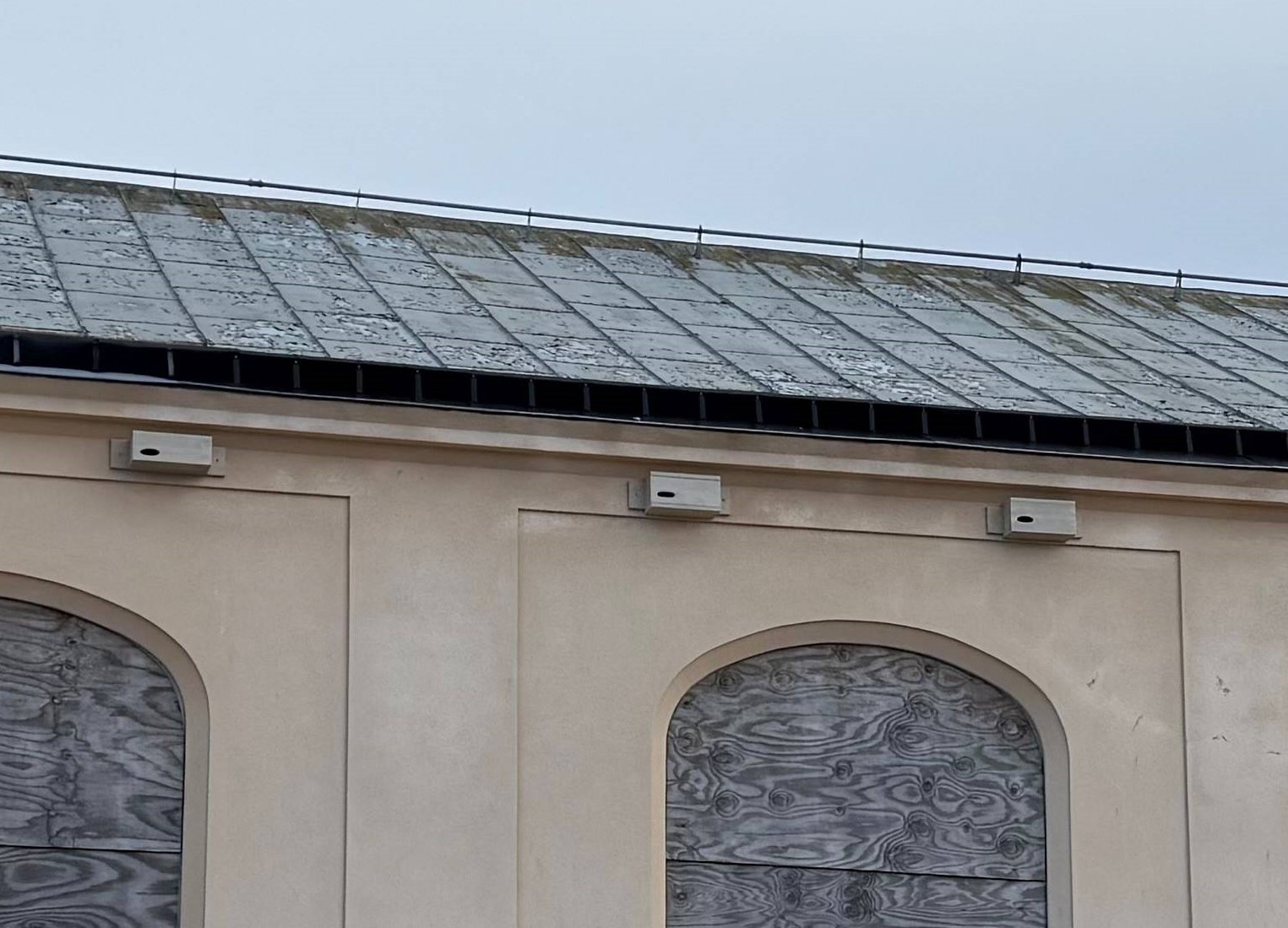 En byggnad med ett snett plåttak och igensatta bågformade fönster. Tre rektangulära fågelholkar sitter på väggen under taket.