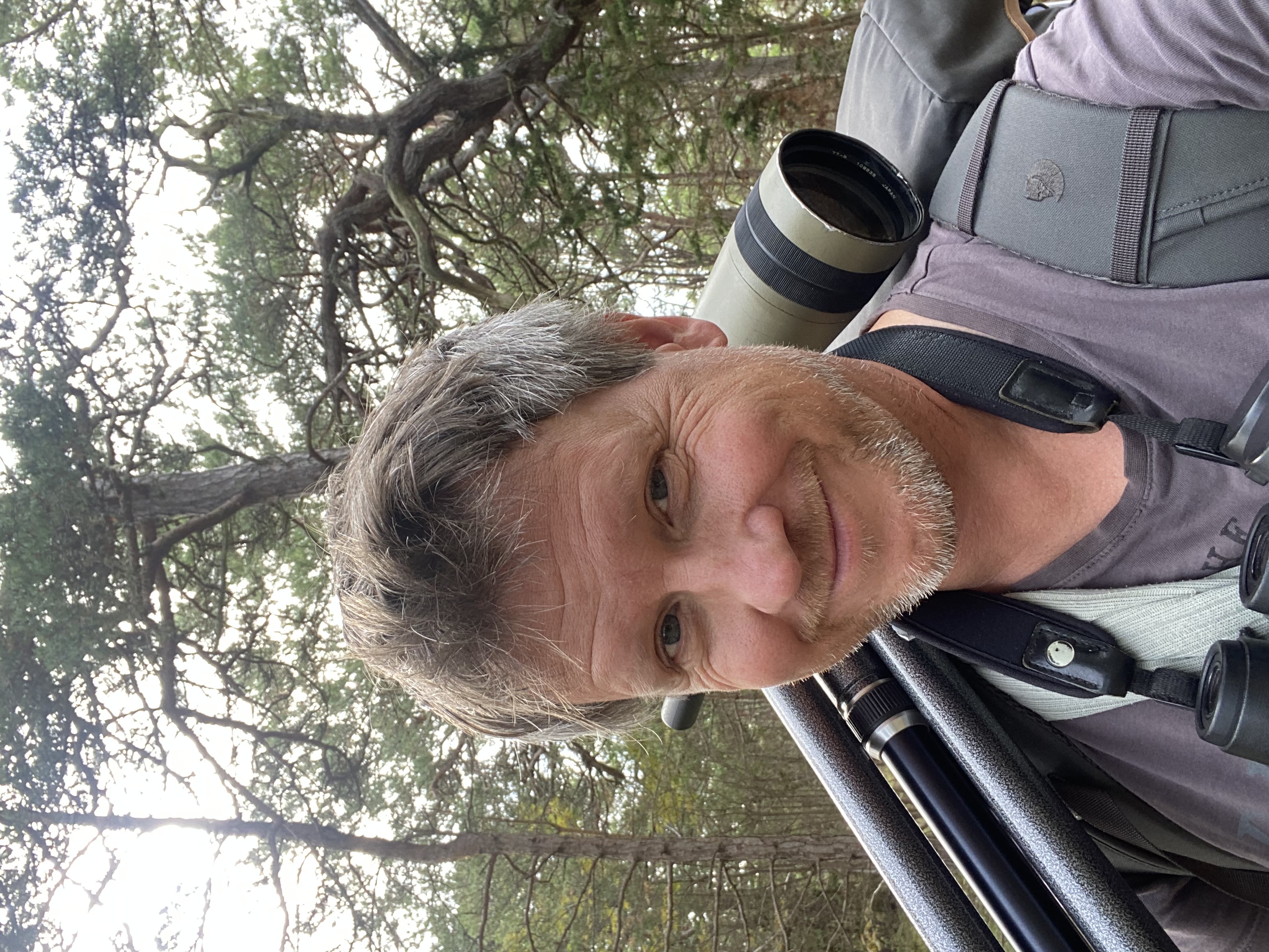 Man med kikare och kamerautrustning ler mot kameran och står utomhus i ett skogsområde.