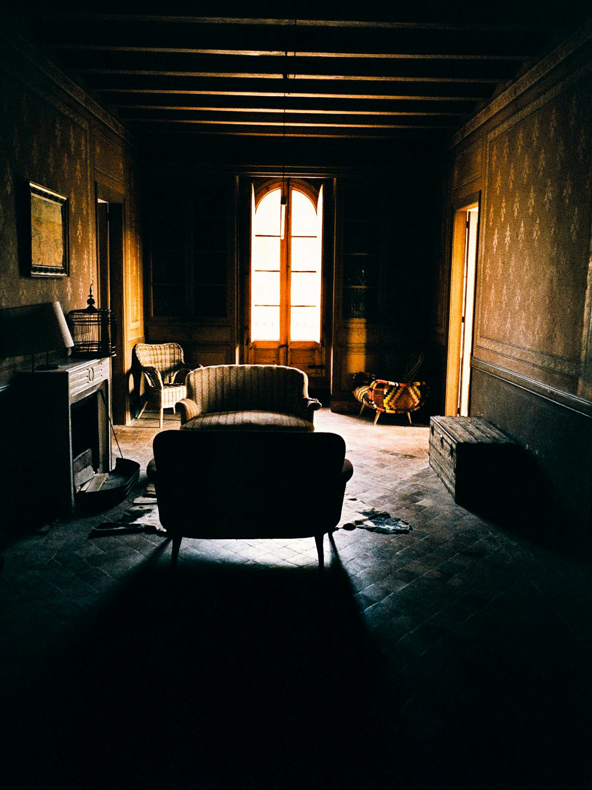 Ett svagt upplyst äldre vardagsrum med en soffa, fåtölj och öppen spis, solljus strömmar genom ett öppet fönster.