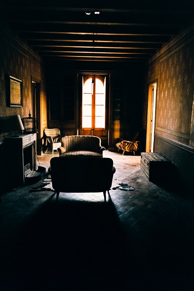 Ett svagt upplyst äldre vardagsrum med en soffa, fåtölj och öppen spis, solljus strömmar genom ett öppet fönster.