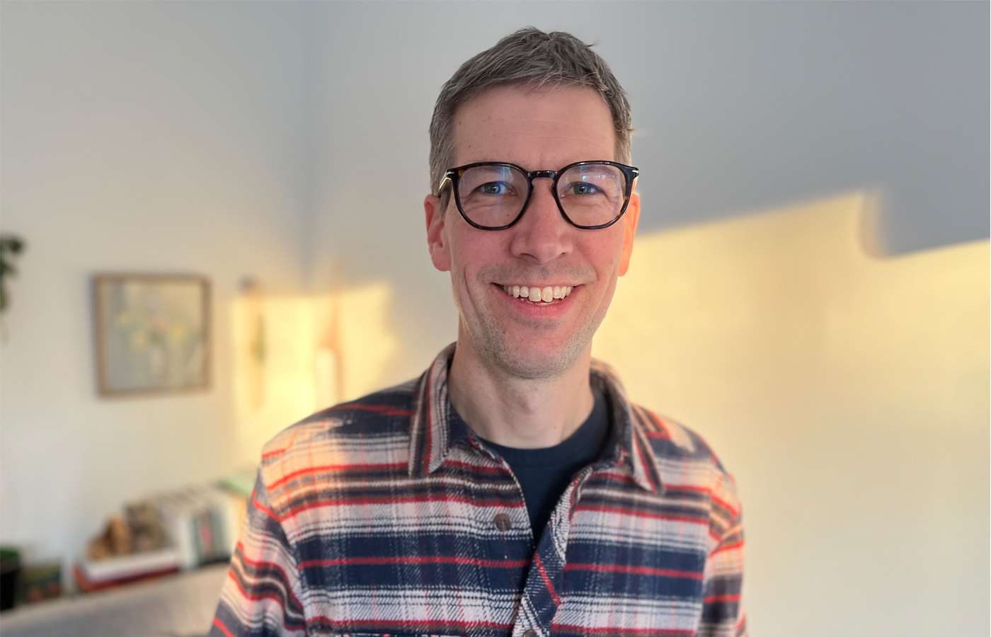 Ola Karlsson ler mot kameran och har en randig skjorta på sig. I bakgrunden syns en tavla i ram och några böcker.