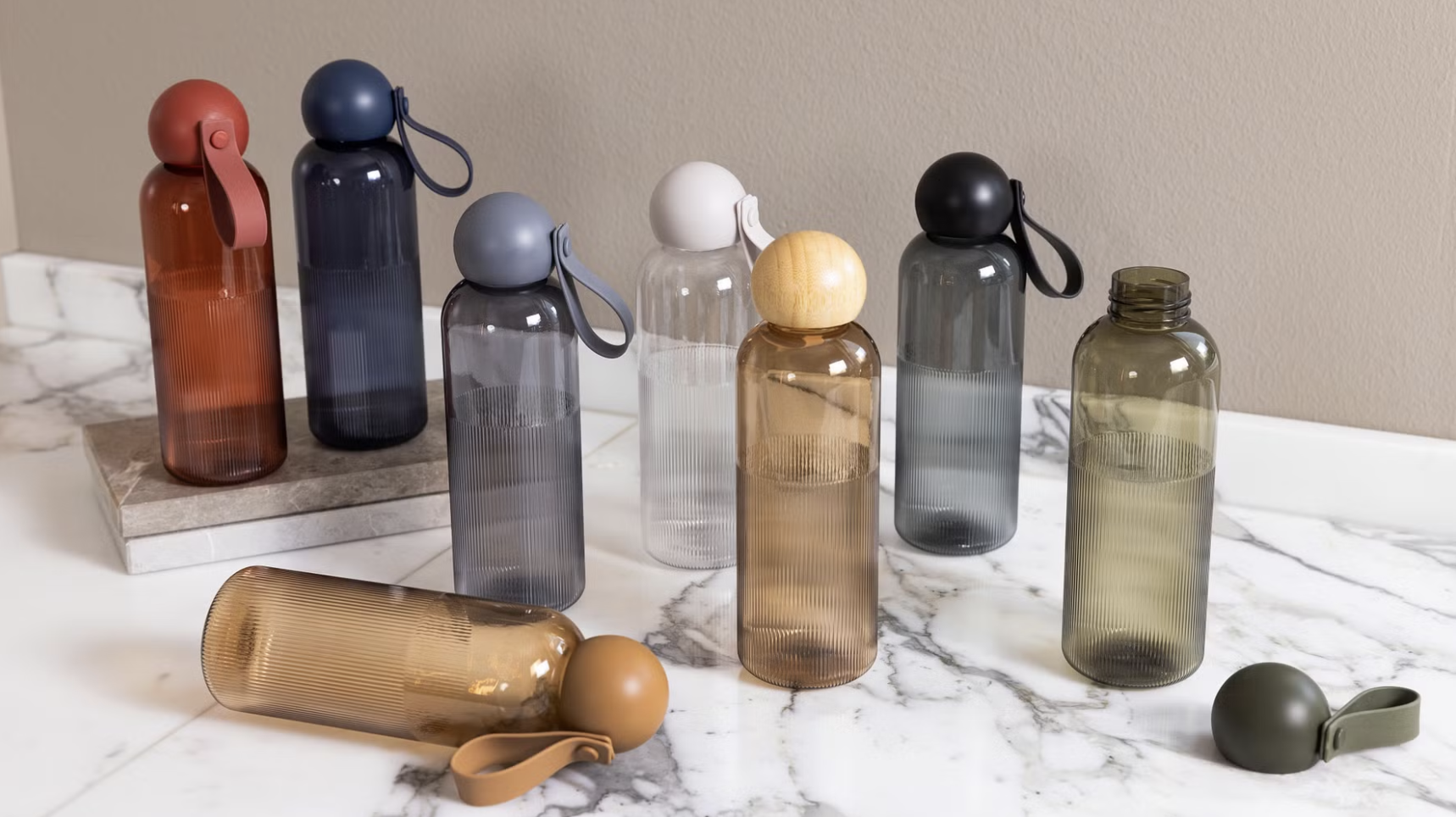 Åtta plastflaskor av transparent material med olika färgade lock och remmar står uppställda på en yta av marmor och stenhällar.