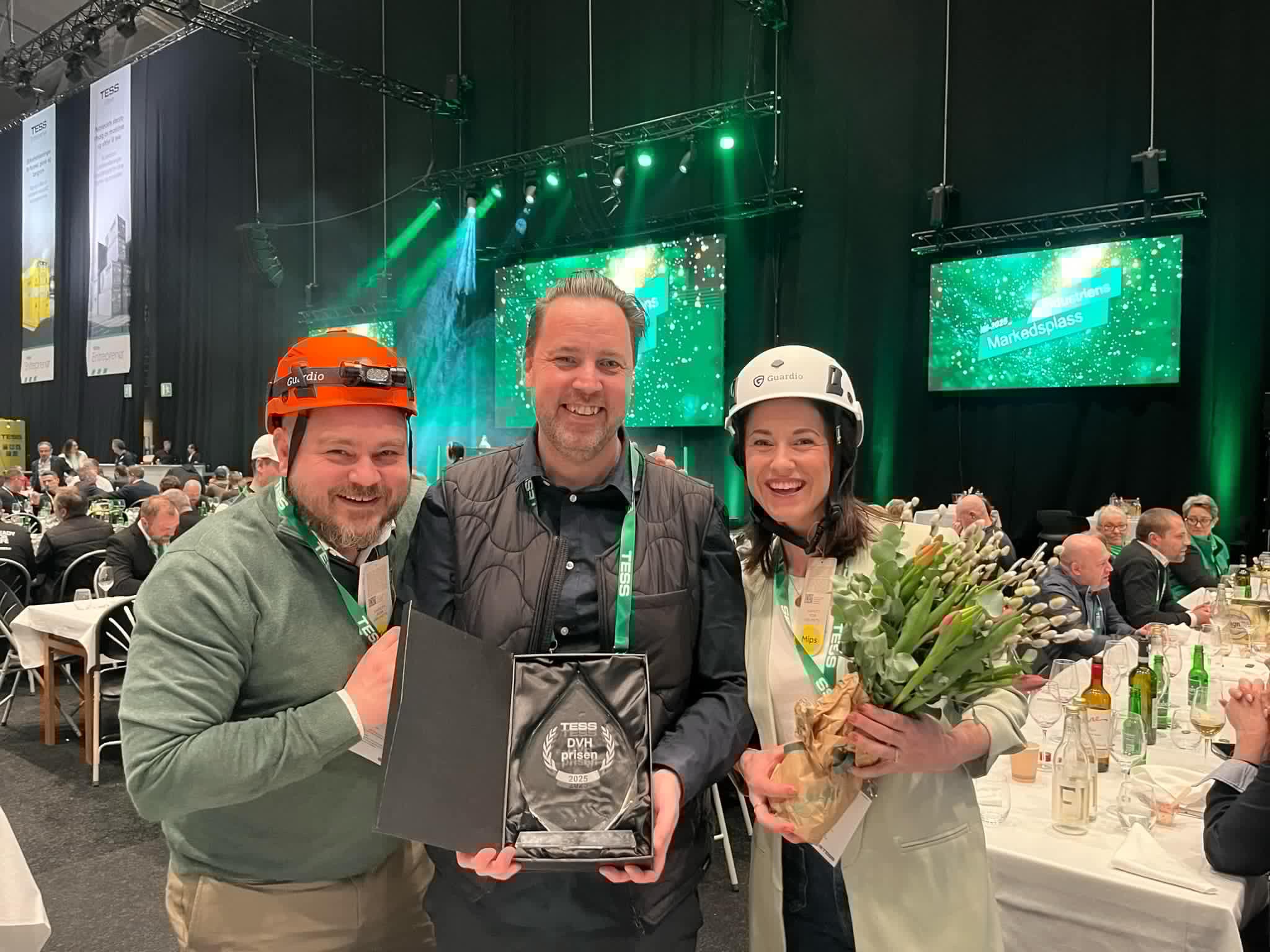 Tre personer med hjälmar står leende vid ett inomhusevenemang. En håller en plakett, och en annan har en bukett. Bakom dem finns en scen upplyst i grönt och publik.
