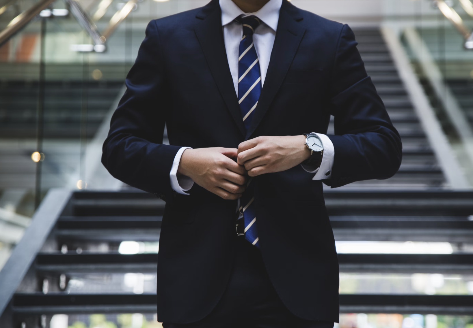 En person i kostym och randig slips knäpper sin kavaj och står framför en trappa i en byggnad.